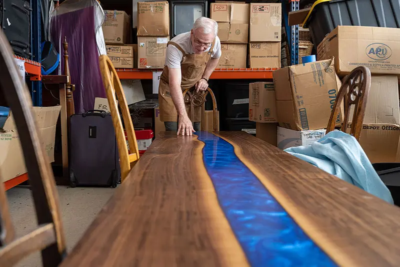 A craftsman wearing a tan apron leans over a long, handcrafted wooden table. A vibrant blue epoxy resin inlay, resembling a river, flows down the center of the table. The surrounding storage area is filled with cardboard boxes, chairs, and a suitcase.