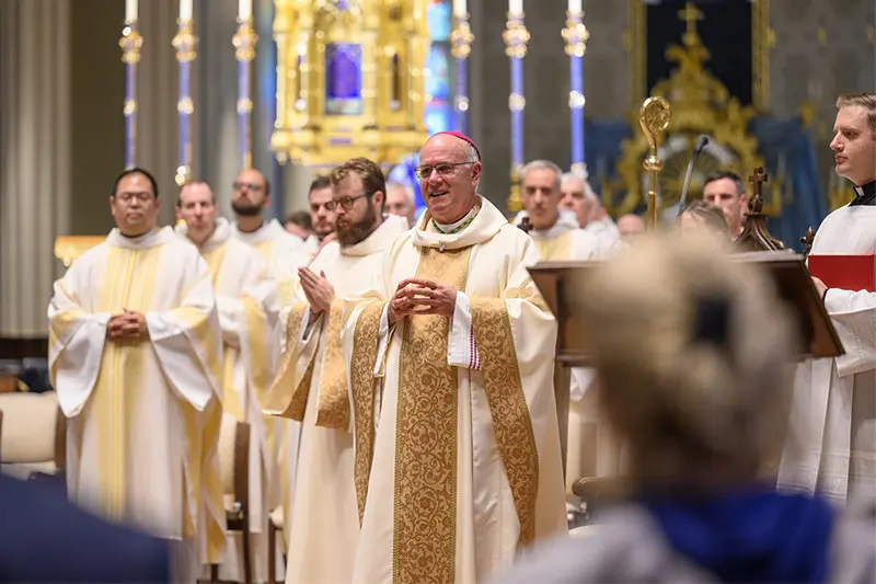 A bishop, wearing a white chasuble with gold embroidery, gestures with his hands while addressing a congregation. Several priests, also in white and gold vestments, stand behind him in the cathedral. A blurred figure in the foreground suggests the perspective of an audience member.