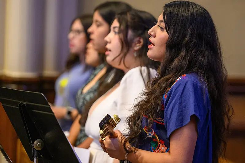 Four women sing, facing left. The woman in the foreground, wearing a blue embroidered top, plays a small stringed instrument. A music stand is partially visible on the left.