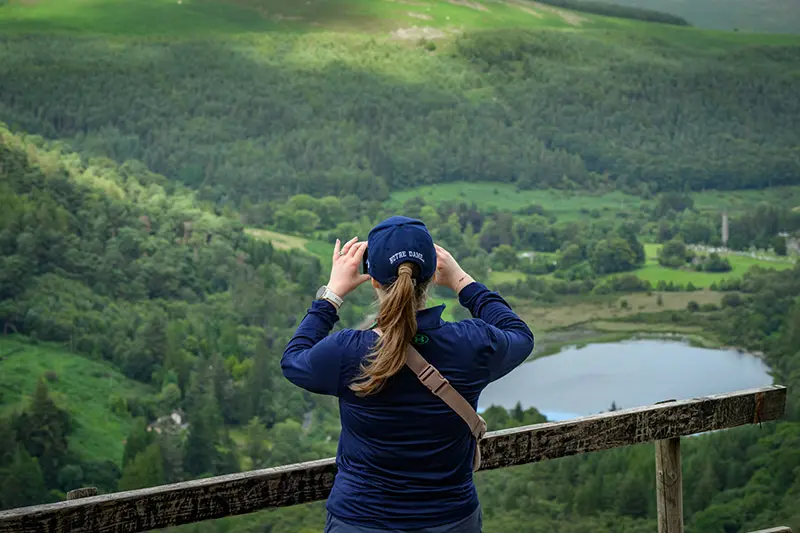 A person wearing a Notre Dame baseball cap takes a photo of a scenic overlook. Lush green hills slope down to a tranquil lake, surrounded by trees. The person stands behind a weathered wooden railing.