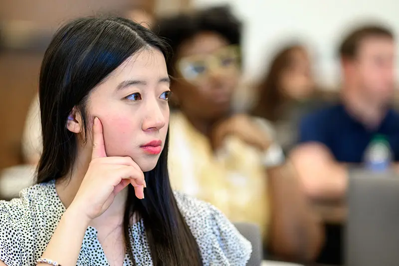 An asian woman with long, dark hair rests her chin thoughtfully on her hand during a class at the University of Notre Dame. Other students are visible but out of focus in the background.