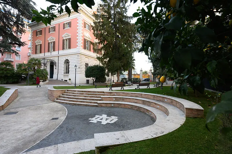 The University of Notre Dame’s Rome Global Gateway. A three-tiered, semi-circular brick amphitheater sits in the foreground with the Notre Dame logo in the center. The pink and white building which houses the Rome Global Gateway is in the background. A few park benches and a lemon tree are visible.