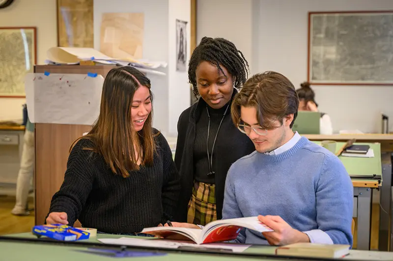 Three students collaborate on a project. Two women lean over a table to look at a book held open by a male student. Maps and architectural sketches hang on the walls in the background.