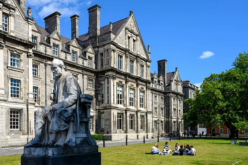 A statue of a seated man in academic robes sits prominently in the foreground of a photo depicting a grand, multi-story stone building on a sunny day. In the background, a group of students sits in a circle on the grassy lawn.