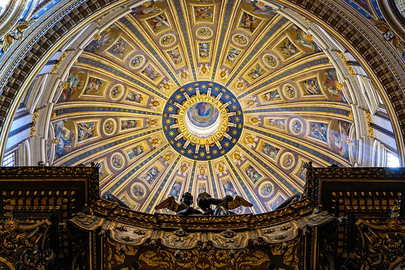 Looking upward at the ornate dome of St. Peter's Basilica. The apex features a circular skylight surrounded by Latin inscriptions and gold stars on a navy background. The rest of the dome is intricately decorated with gold ribs, mosaics of religious figures, and detailed gold and blue accents. The edge of an ornate wooden structure is visible in the foreground.