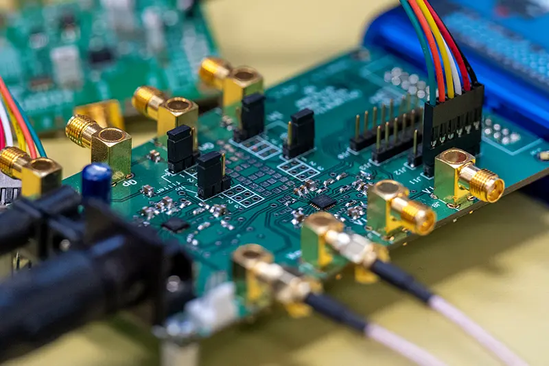 Close-up of a green circuit board. Gold connectors are attached to the board, along with black components and a multi-colored wire connector. Other circuitry is visible in the blurred background.