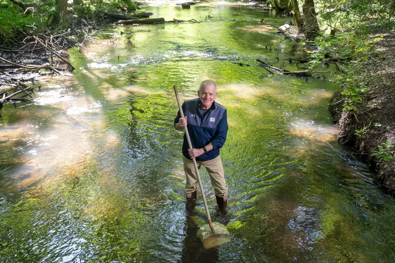 A man wearing a Notre Dame jacket and khaki pants stands in a shallow, clear stream. He holds a long pole, its end resting on a submerged rock, likely for stability. Sunlight dapples the streambed through the trees lining the banks.