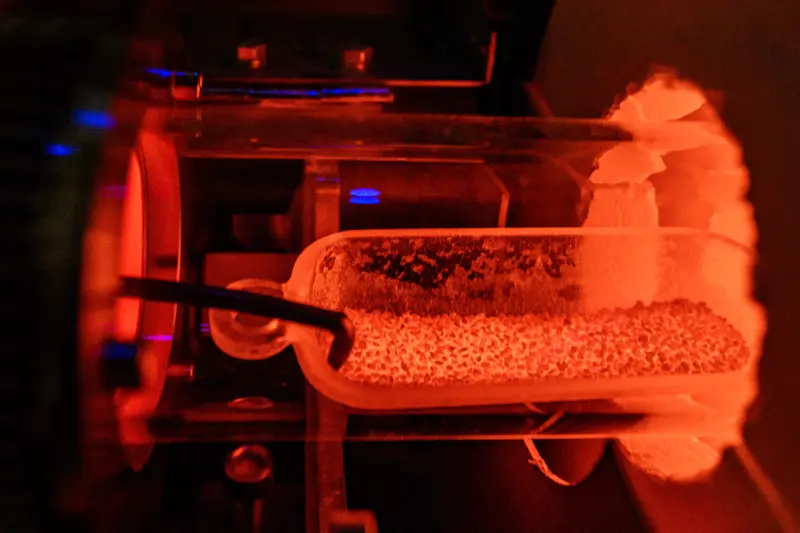 A small, clear, rectangular container filled with a grainy substance glows under orange light within a larger piece of scientific equipment. A thin metal rod extends into the container.