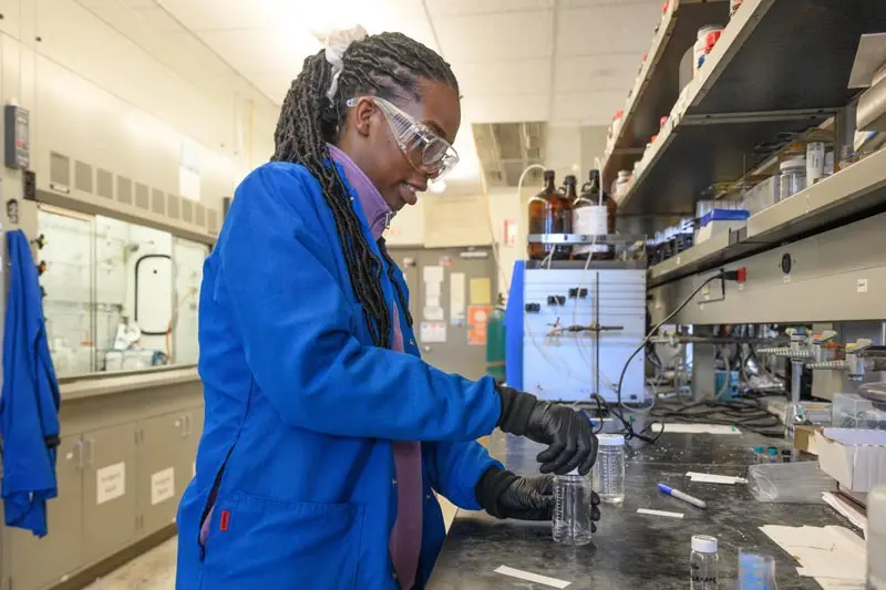 A black woman wearing a blue lab coat, safety glasses, and black gloves carefully handles clear containers filled with liquid at a lab bench. Various lab equipment, including bottles, containers, and instruments, are visible on shelves and the counter in the background.