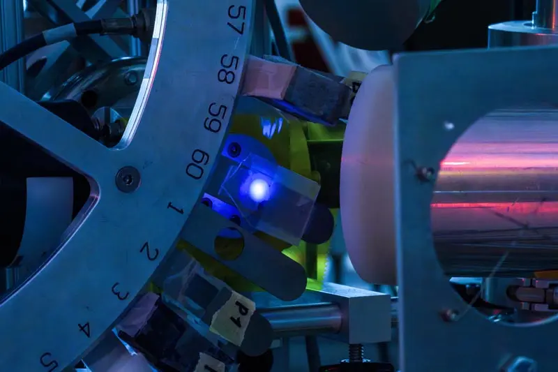 Close-up of a scientific instrument. A circular dial numbered 57 through 4 is partially visible on the left. A bright blue light emanates from a component near the dial's center. This light is directed towards a cylindrical object held within a metal frame. A reddish glow is visible within the cylinder. The scene is bathed in a cool, bluish light.