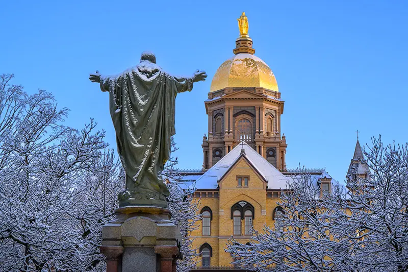 A statue of Jesus with outstretched arms, dusted with snow, stands before the iconic golden dome and snow-covered roof of the Main Building at the University of Notre Dame. Bare tree branches, also coated in snow, frame the scene against a clear blue sky.