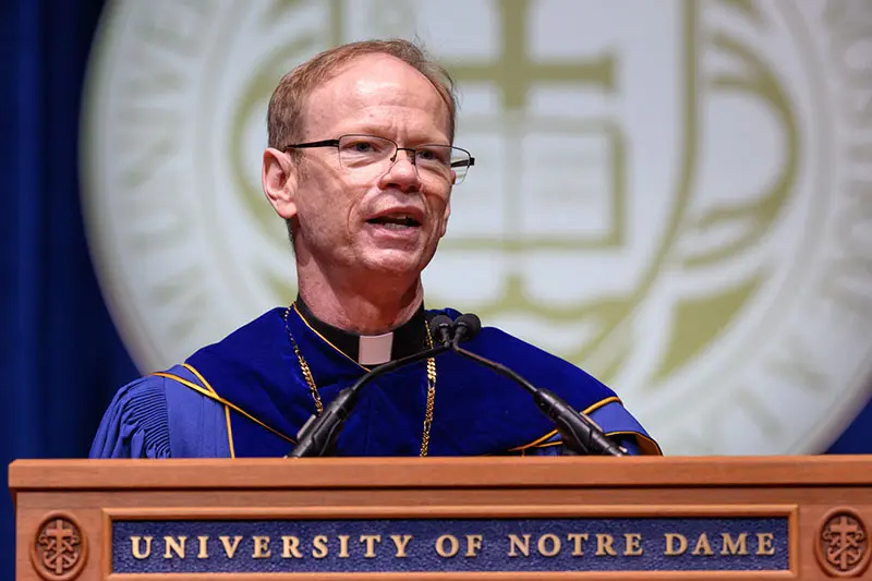 A man wearing glasses and academic regalia speaks at a podium. The podium has UNIVERSITY OF NOTRE DAME engraved on the front. A blurred image of the University of Notre Dame seal hangs in the background.