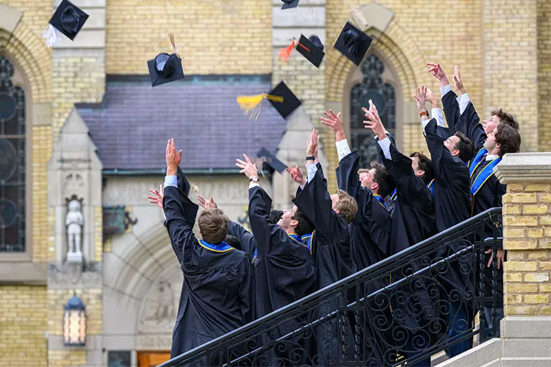 Graduates in black gowns and gold-trimmed blue stoles toss their mortarboards into the air in front of a light-colored stone building. They stand on steps with a decorative black railing.