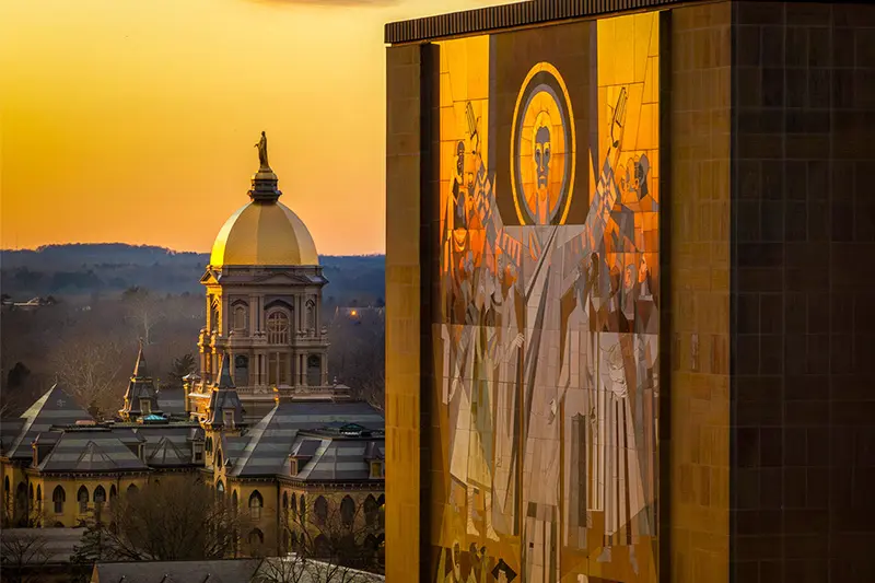 At sunset, the iconic golden dome of the Main Building at the University of Notre Dame is seen in the background. In the foreground, a section of the large mosaic known as The Word of Life is displayed on the Hesburgh Library.