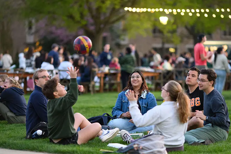 Students gather on a grassy quad at an evening event. String lights twinkle in the background. In the foreground, a small group sits on the grass, one tossing a volleyball in the air, while others watch and gesture.  Additional students and picnic tables are visible in the background.