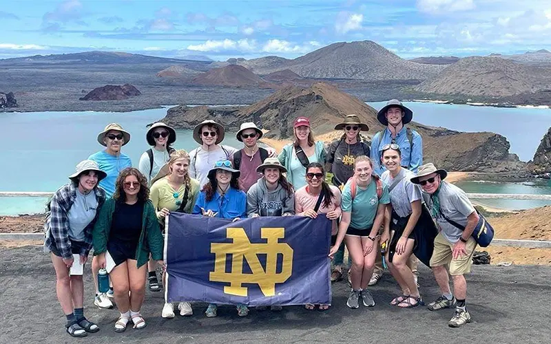 A group of University of Notre Dame students and faculty pose with a large Notre Dame flag in the Galapagos Islands. They stand on a volcanic overlook with a stunning view of turquoise waters, small islands and volcanic peaks.