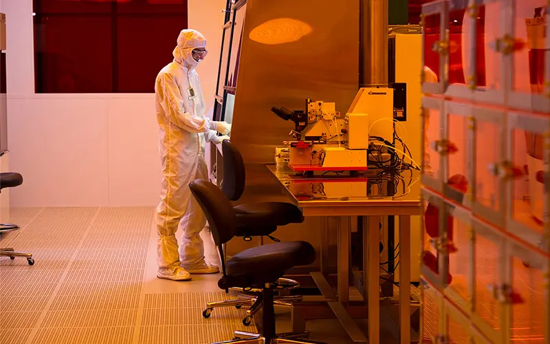 A researcher in a white clean suit operates a microscope in a brightly lit laboratory. The room has a perforated orange floor, and orange light reflects off the stainless steel surfaces. To the right, a stacked structure with transparent orange panels stores lab materials. Two rolling chairs sit in front of the workstation.