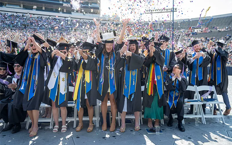 Architecture students from Notre Dame's Class of 2024 toss confetti into the air at the conclusion of their commencement ceremony in Notre Dame Stadium. They are wearing graduation caps and gowns.