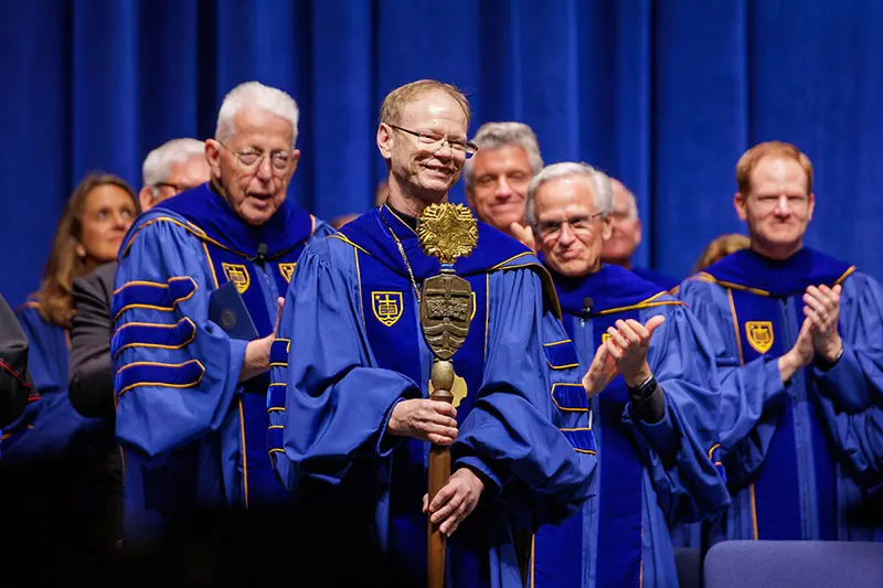 Father Dowd standing in his academic regalia and holding the University Mace.