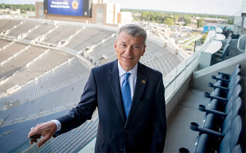 A white man with a white shirt, blue tie, and navy suit coat is standing in the stands of Notre Dame stadium.