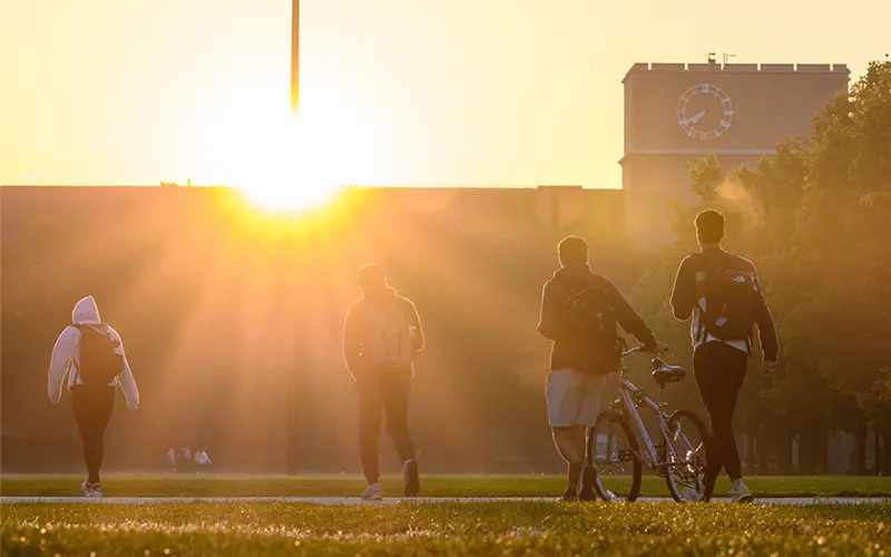 A group of students walk in the direction of an an academic building silhouetted by the sun.