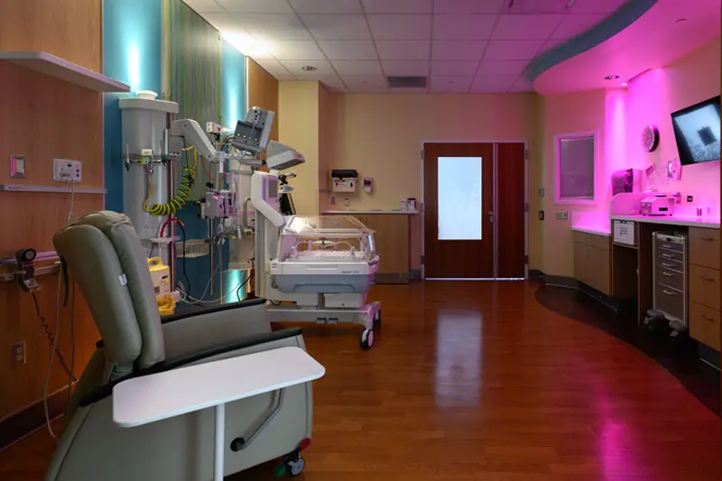 A private hospital room designed for postpartum care. In the foreground, a reclining chair with a pull-out table faces a hospital bassinet and medical equipment. Soft, colorful lighting illuminates the teal and pink walls, creating a calming atmosphere. A closed double door is visible in the background.