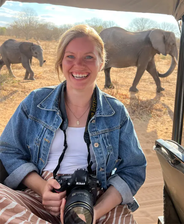 Grace Clinton sits in an open vehicle holding a camera. There are elephants in the background.