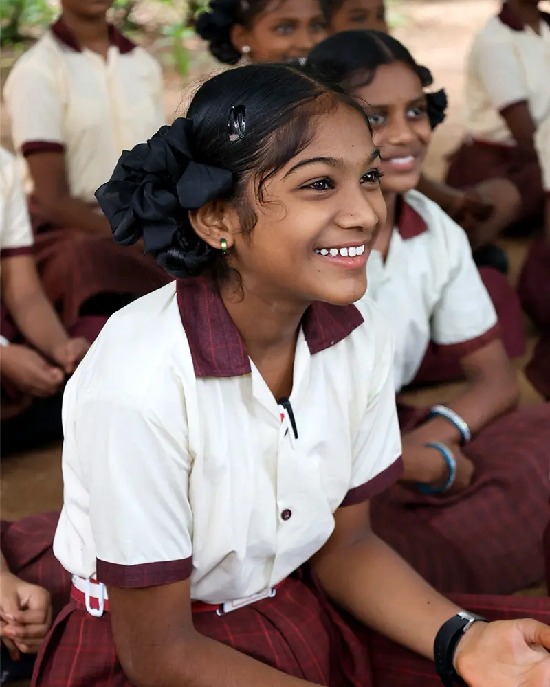 A young Indian girl with a large black hair bow smiles brightly while sitting with other students. She wears a white and burgundy school uniform.