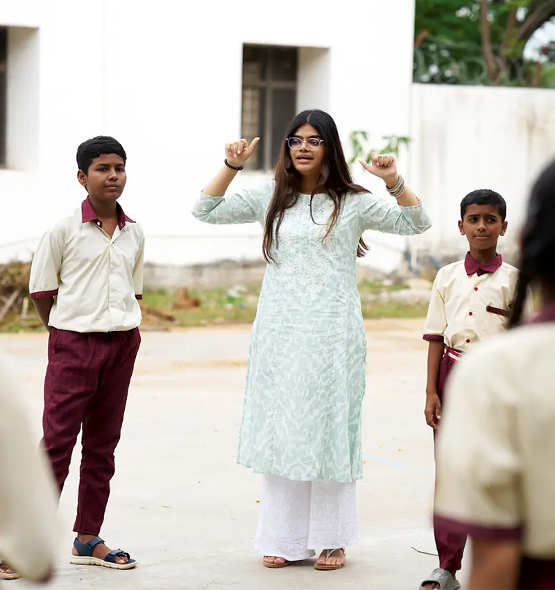 A brown-skinned woman in a light green and white outfit gestures with her hands while leading an activity for two children in school uniforms on a paved area.