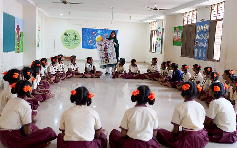 A white woman holds a poster depicting a tree with figures on its branches while teaching a class of young girls sitting in a circle on the floor. Colorful educational posters decorate the walls of the classroom. The Indian girls all wear matching cream shirts and maroon skirts and have orange flowers in their hair.