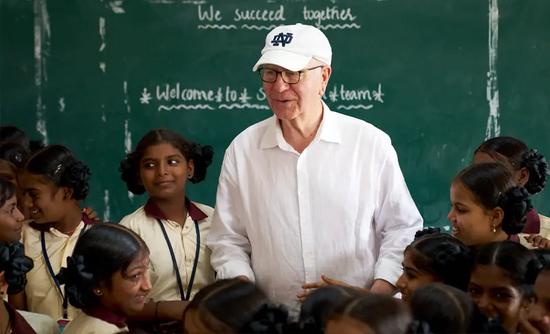 A white man in a white Notre Dame baseball cap and shirt smiles at a classroom of young Indian girls in uniform in front of a chalkboard that reads 