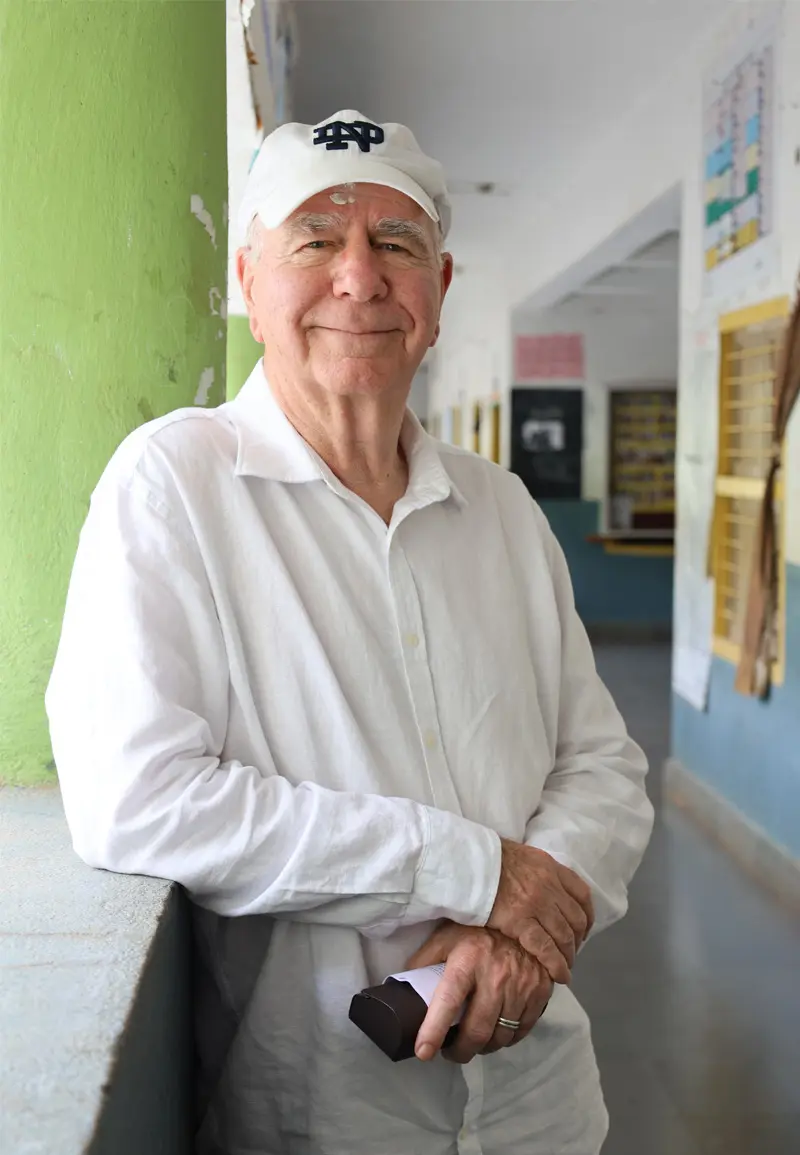 An older white man wearing a white Notre Dame baseball cap and a white, long-sleeved shirt stands in the hallway of a school building. He leans against a window sill and smiles toward the camera. 