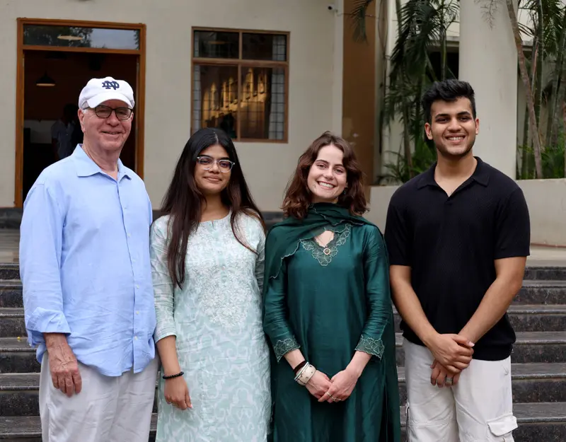 Four individuals stand smiling in front of a light-colored building with stairs. From left to right: a white man in a light blue shirt and Notre Dame baseball cap, a brown-skinned woman in a light teal patterned kurta, a white woman in an emerald green salwar kameez, and a brown-skinned man in a black polo shirt and khaki pants.