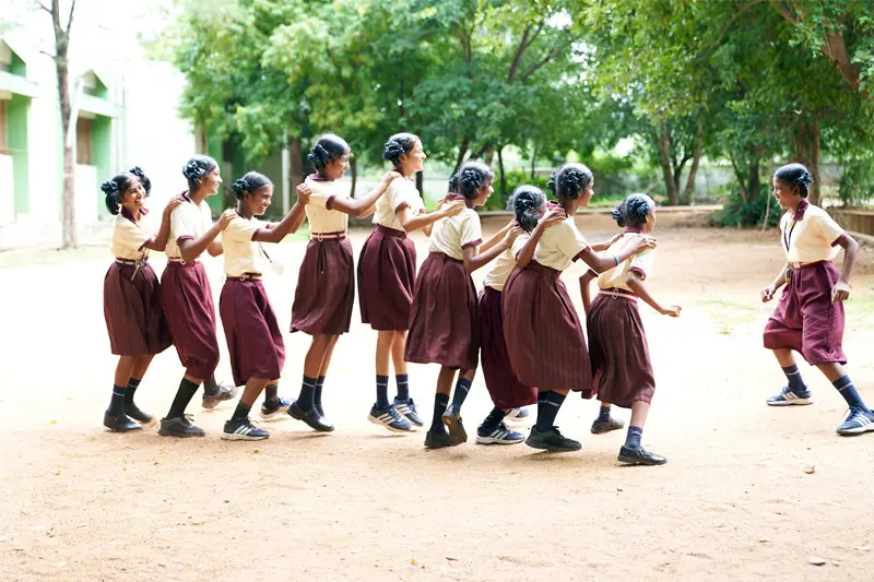 A line of Indiana girls in matching tan and maroon school uniforms hold each other's shoulders and playfully skip across a dirt schoolyard. Trees and a school building can be seen in the background.