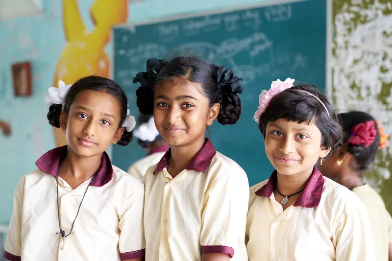 Three young Indian girls in cream and maroon school uniforms stand smiling in front of a teal chalkboard in a classroom. Two girls wear white flowers in their hair, while the third wears a pink flower headband.