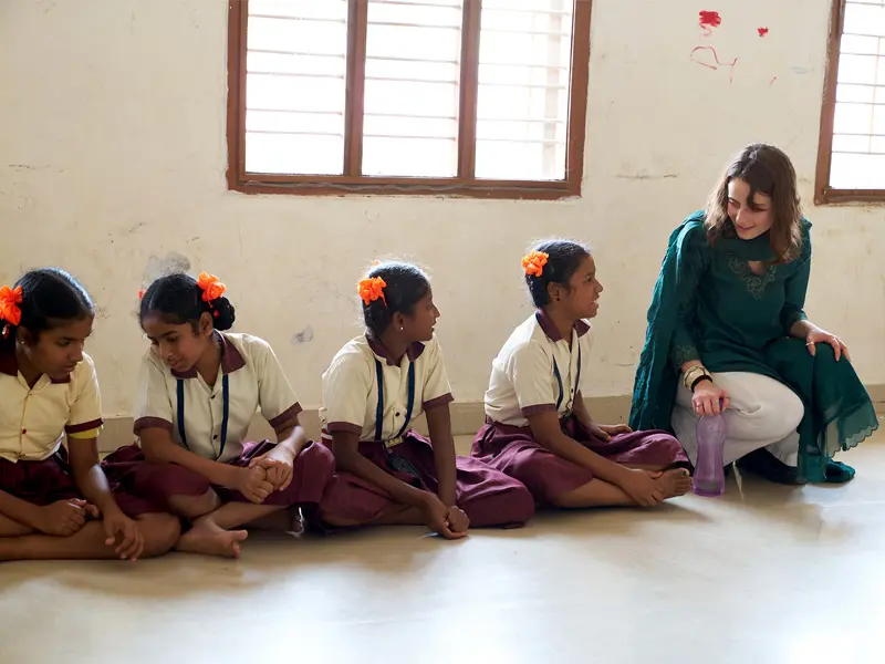 Four girls in school uniforms sit on the floor facing a woman kneeling beside them. The girls have orange flowers in their hair and wear tan and maroon uniforms. The woman wears a green kurta and white pants. 