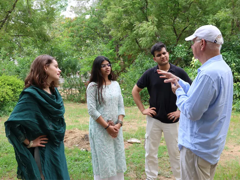 Four individuals stand in a grassy area surrounded by trees. Two women in traditional South Asian attire and a man wearing a black polo shirt and khaki pants listen as an older man wearing a light blue shirt, khaki pants, and a white baseball cap gestures while speaking to them.