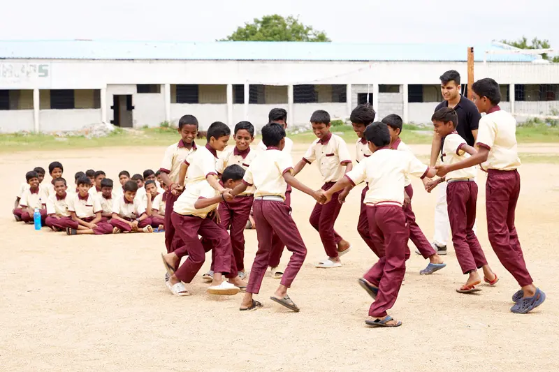 Indian schoolchildren in tan and maroon uniforms hold hands and joyfully dance in a circle on a dirt play area. Another group of children sit and watch in the background. A young man in a black shirt and white pants stands near the dancing children. A partially visible building stands in the background.