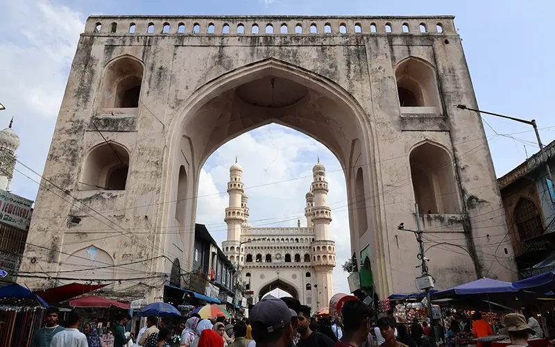 A large arched gateway, weathered and pale, dominates the foreground. Through its opening, the Charminar, a grand mosque with four towering minarets, is visible. A bustling street market stretches from the gateway towards the Charminar, filled with people and colorful vendor stalls. 