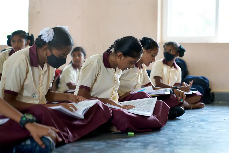 A group of young Indian girls in matching cream and maroon uniforms sit cross-legged on a classroom floor engrossed in their textbooks.