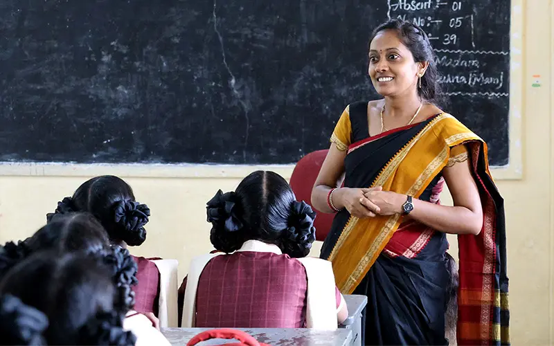 A brown-skinned teacher in a sari smiles warmly while facing a classroom of young girls in India. The girls, seen from behind, wear maroon and white uniforms and have their dark hair styled in buns. A chalkboard hangs on the wall behind the teacher. 