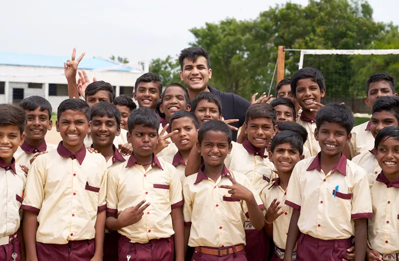 A group of smiling children in matching khaki and burgundy school uniforms pose with a young man in a black shirt. Several students hold up peace signs, adding to the cheerful atmosphere. 