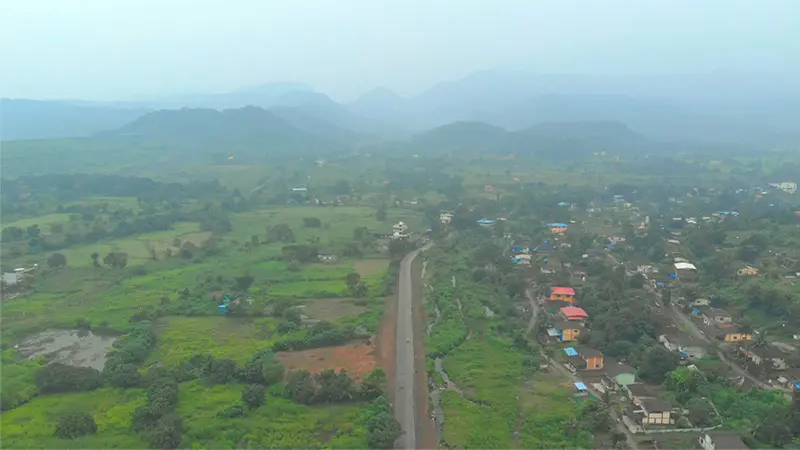 Aerial view of a rural landscape with a two-lane road cutting through verdant fields and a small village nestled among trees. Misty mountains rise in the background.