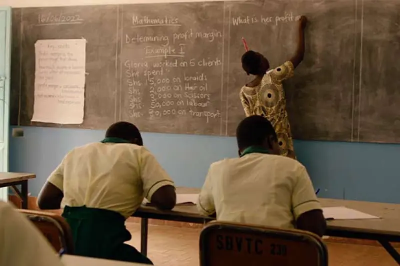 Students in cream-colored shirts and teal pants sit at desks working, their backs to the camera. A partially visible teacher writes a mathematics problem on determining profit margin on a chalkboard at the front of the class. A piece of paper is also taped to the chalkboard.