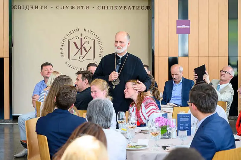 A group of people sit at a table during a formal dinner or reception. A priest stands and speaks, gently placing his hand on a woman's head. The Ukrainian Catholic University logo is visible on the wall behind them.