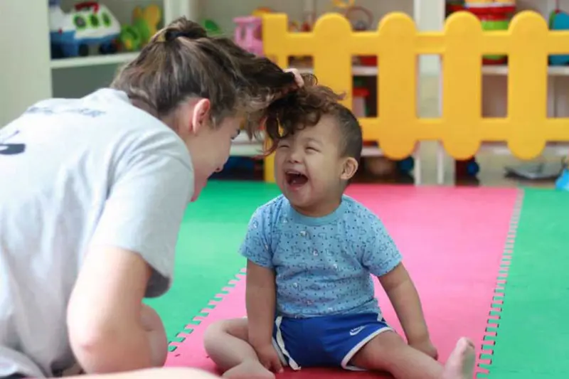 A Notre Dame student leans toward a laughing toddler sitting on a colorful play mat. The student playfully holds the toddler's hair. Toys and a yellow fence are visible in the background.