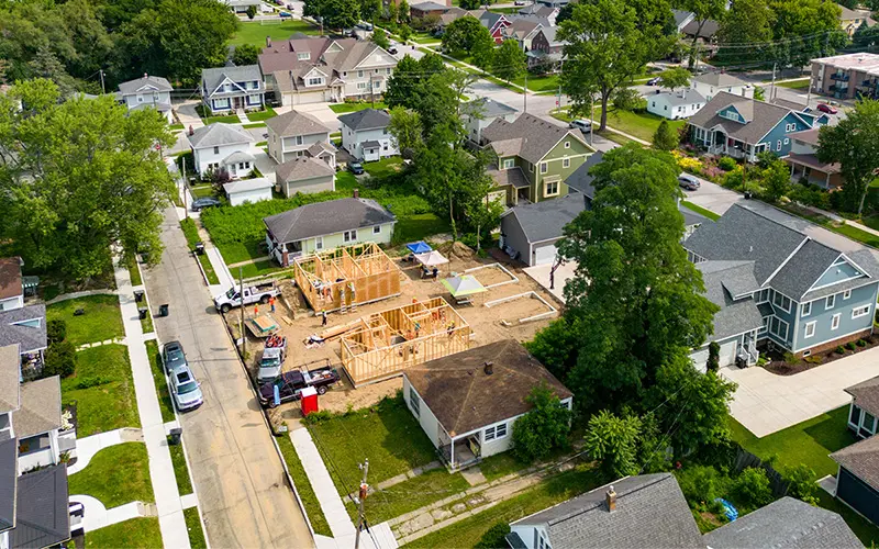 Aerial view of a residential neighborhood with two homes under construction. The wooden frames of the houses are visible, and construction workers are on site. Trucks and equipment are parked nearby. Surrounding the construction site are established homes with yards and driveways.