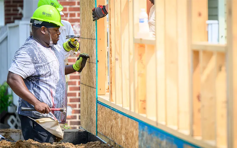 A construction worker wearing a lime green Habitat for Humanity hardhat uses a hammer to build the frame of a house.  He's wearing work gloves and holding the hammer in his left hand.