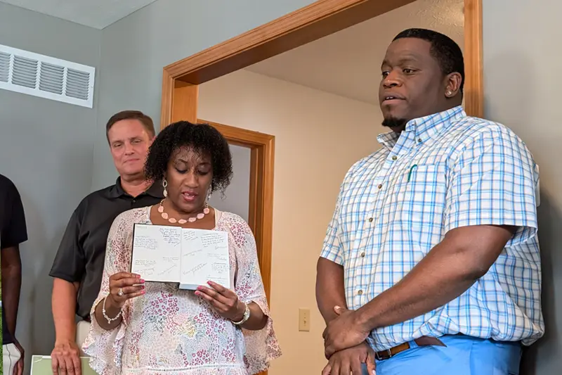 A woman reads aloud from a notebook filled with handwritten messages.  A man stands listening behind her to her left, and another man stands to her right with his hands clasped. They appear to be inside a house or office.