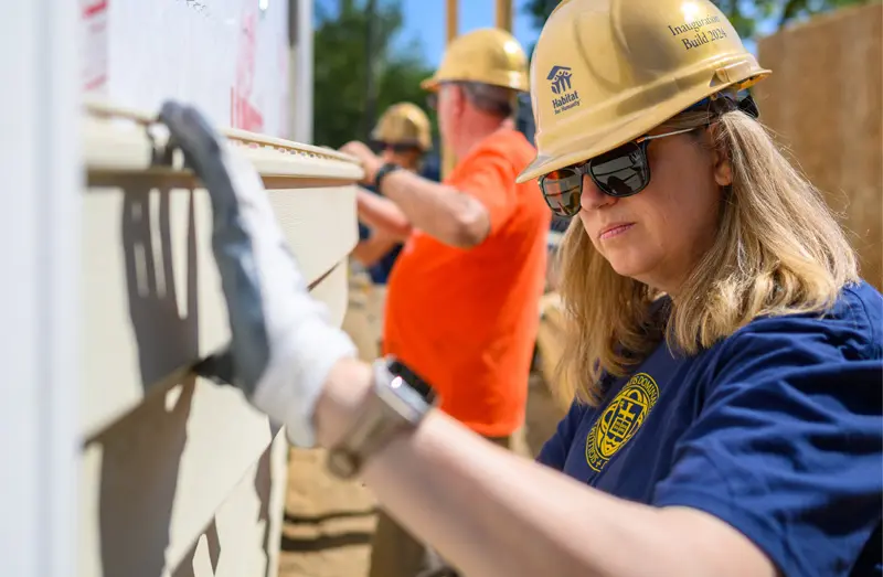 A woman wearing a gold hardhat, sunglasses, and a University of Notre Dame t-shirt installs siding on a Habitat for Humanity house. Other volunteers in hard hats work in the background.